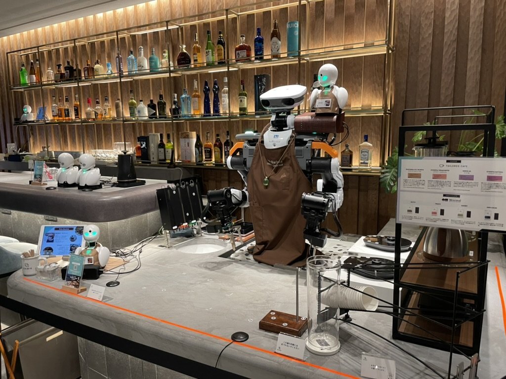 A remotely operated robot at the café counter, with bottles displayed on shelves behind it